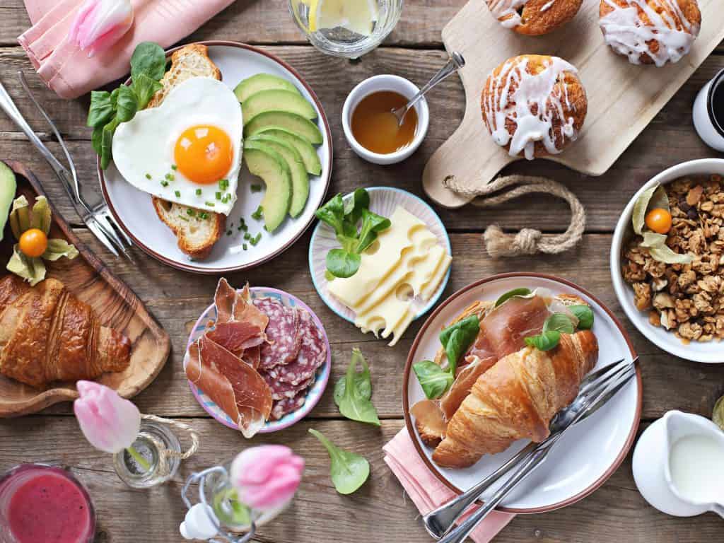 Breakfast buffet seen from above on a rustic wooden table: fried eggs with avocado, croissants with ham, a cheese and cold-cut platter, granola, muffins, and honey, along with tulips and a berry smoothie.